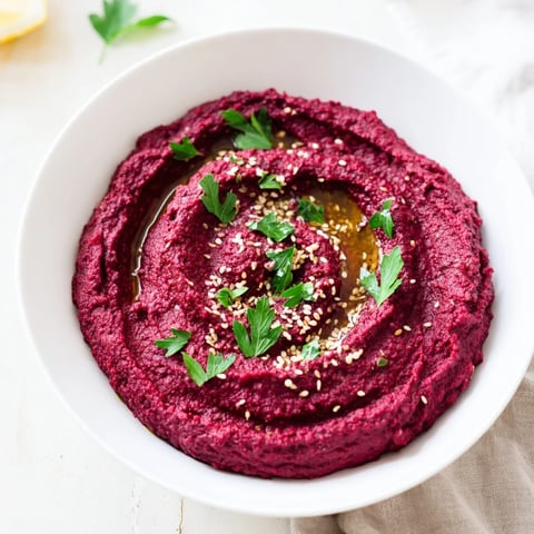 Vibrant roasted beet hummus in a white bowl, garnished with parsley and a drizzle of olive oil, perfect for dipping fresh vegetables.  