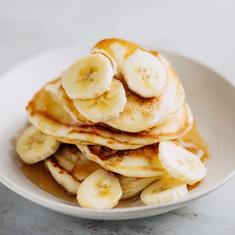 Stack of golden, cooked Weekend Golden-Brunch Banana Pancakes, drizzled with sweet syrup and sliced bananas.